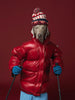 a photograph by William Wegman of a weimaraner wearing a red puff jacket and a knitted hat